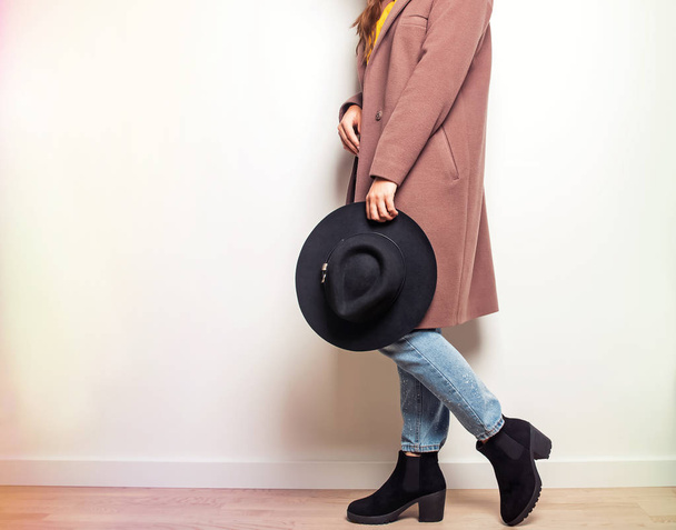 Женщина в бежевом пальто и стильных сапогах с шляпой в руке
. - Фото, изображение