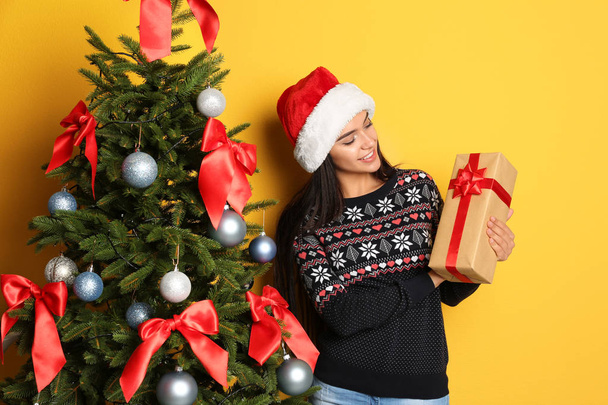 Beautiful young woman in Santa hat with gift box near Christmas tree on color background - Fotó, kép