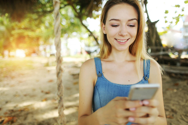 Sunshine ritratto di giovane ragazza bionda che chiacchiera con lo smartphone e l'altalena a Bali, sabbia sullo sfondo
. - Foto, immagini