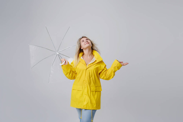 Blonde vrouw draagt gele regenjas holding transparante paraplu controleren weer als het regent. Beschermd tegen regen en orkaan - Foto, afbeelding