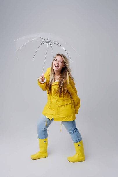 Blond žena nosí žluté pláštěnce drží transparentní deštník kontrolu počasí, pokud prší. Chráněno před deštěm a hurikán - Fotografie, Obrázek