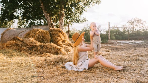 woman in hat and cute little boy in linen clothing resting on hay at countryside - Foto, Imagem