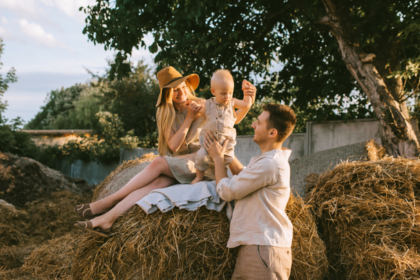 family with little son in linen clothing resting on hay at countryside - Foto, afbeelding