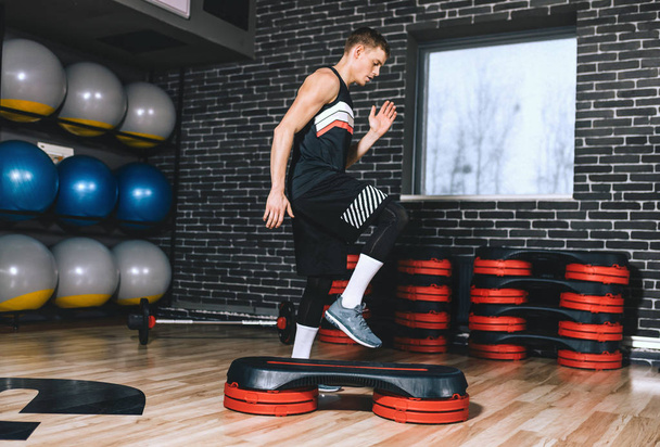 Side full body view of sporty athlete man doing step up jumps in health fitness club. The handsome male is exercising his legs on the stepper in the modern gym. Sport, lifestyle and people concept. - 写真・画像