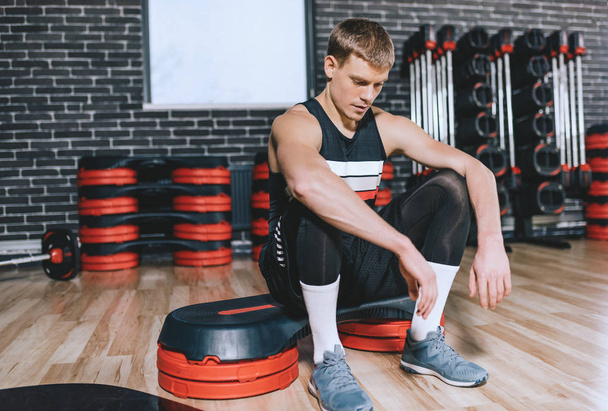 Candid shot of sporty athlete man sitting on stepper relaxing after exercises in the gym. Fitness trainer male rest after hard workouton stepper with copy space for your text or advertising. - 写真・画像