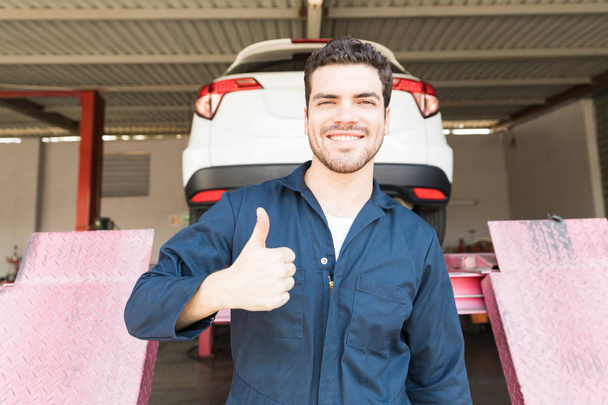 Επιτυχημένη αυτοκινήτων τεχνικός δείχνει τους αντίχειρες επάνω ενώ στέκεται ενάντια αυτοκίνητο ανεβάζω στο κατάστημα - Φωτογραφία, εικόνα