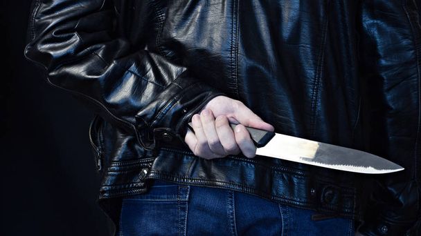 A man in a leather jacket holds a knife behind his back, a black background danger - Φωτογραφία, εικόνα
