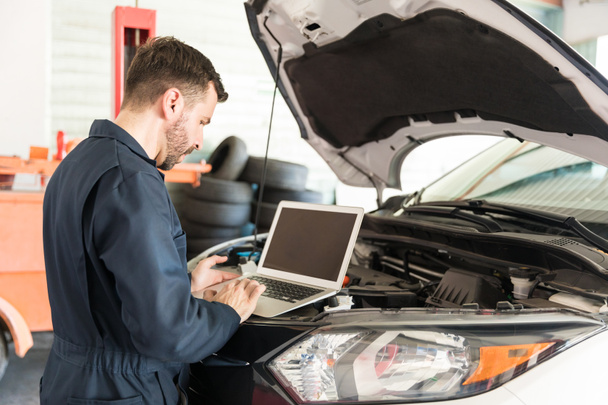 Средний взрослый работник авторемонта, использующий ноутбук для обнаружения неисправности в автомобиле в гараже
 - Фото, изображение