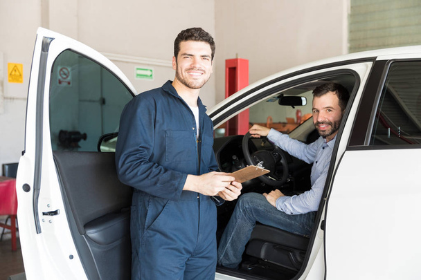 Улыбающийся мужчина-механик держит планшет, стоя рядом с клиентом в машине в гараже
 - Фото, изображение