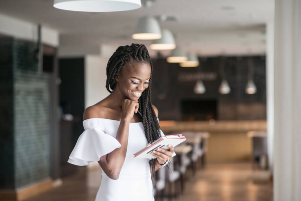 Attracrive african american girl standing in a restaurant or cafe with the tablet in her hands. Dreadlocks or african braids. - Photo, Image