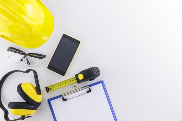 Construction safety wear and tools including hard day, goggles, earmuffs, tape measure, and clipboard plus a mobile phone on white background with copy space - Foto, Bild