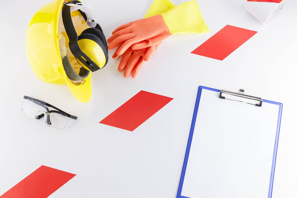 Red and white barricade tape strip separating construction safety gear including goggles, earmuffs, hard hat, and a pair of rubber gloves from a clipboard with blank sheet on white background - Photo, Image