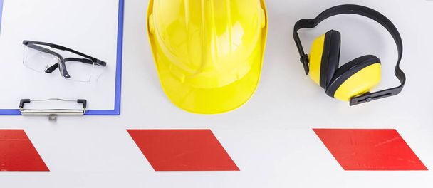 Clipboard e desgaste de segurança de construção, incluindo óculos, chapéu duro e auriculares sobre uma faixa de fita de barreira vermelha e branca na imagem de banner de fundo branco
 - Foto, Imagem