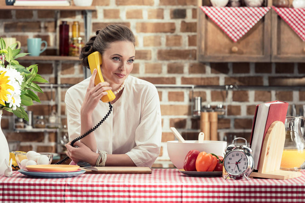 souriant adulte ménagère parler par téléphone filaire rétro et regarder loin de la cuisine
 - Photo, image
