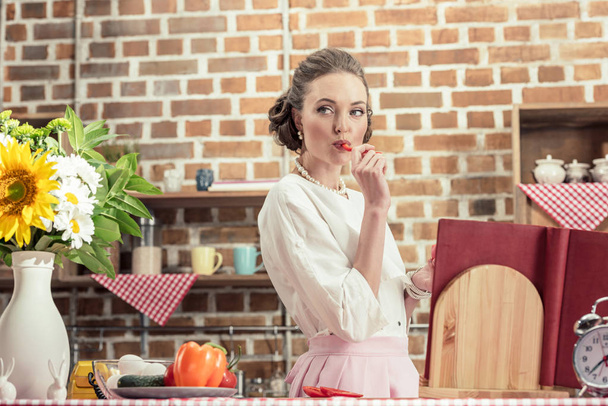 beautiful adult housewife eating raw bell pepper an looking away at kitchen - Photo, Image