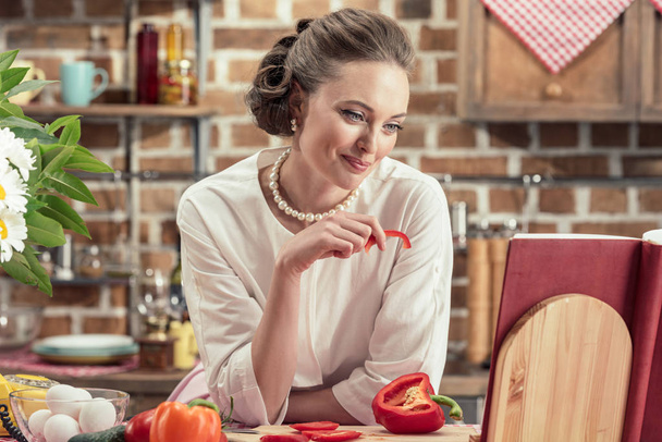 sorridente casalinga adulta che legge il libro di ricette e assaggia verdure fresche in cucina
 - Foto, immagini