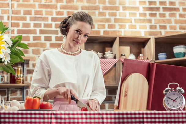 sonriente ama de casa adulta en ropa vintage corte de tomate y mirando a la cámara en la cocina
 - Foto, imagen