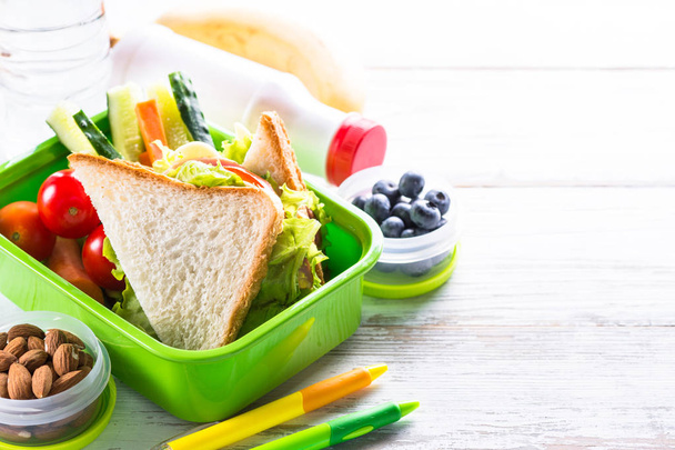Boîte à lunch scolaire avec sandwich, légumes, banane, eau, yaourt, noix et baies sur table en bois blanc. Déjeuner sain à emporter
. - Photo, image