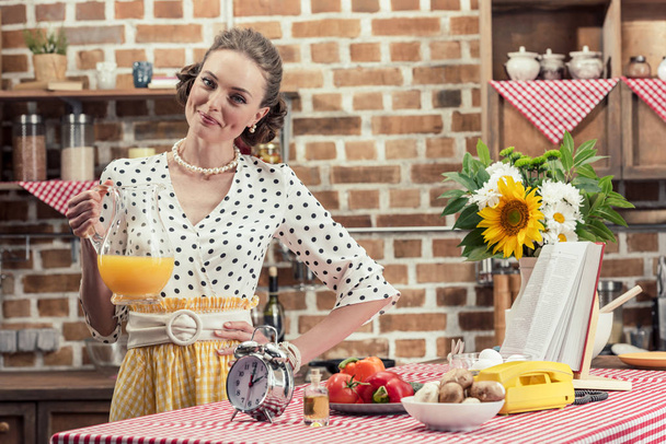 happy adult housewife holding jug of orange juice and looking at camera at kitchen - Fotografie, Obrázek