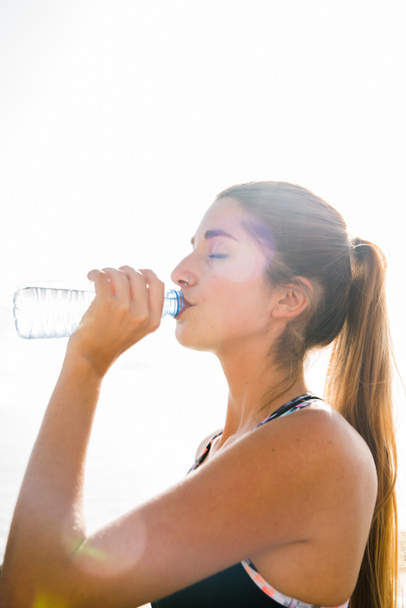 Pony tailed woman is drinking water from a bottle as she closes her eyes and lifts her head towards the sunlight - Photo, Image