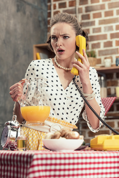 confused adult housewife talking by vintage wired phone at kitchen - Fotó, kép