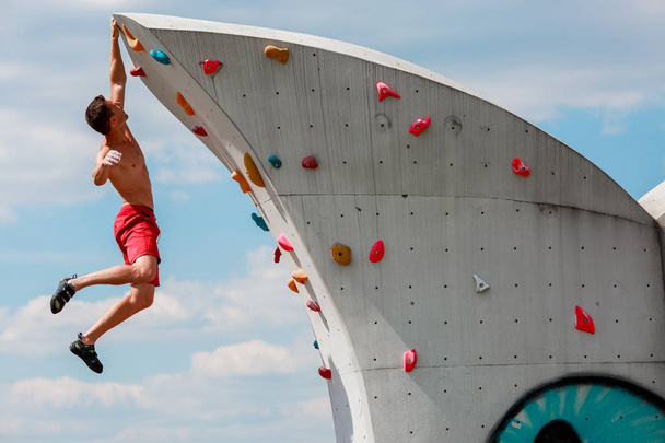 Photo of strong sports man in red shorts hanging on wall for climbing against blue sky with clouds - Zdjęcie, obraz