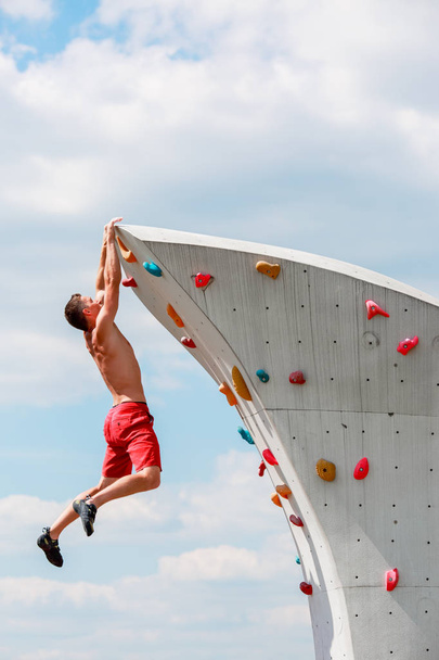 Photo of young sporty man in red shorts hanging on wall for climbing against blue sky with clouds - Фото, зображення