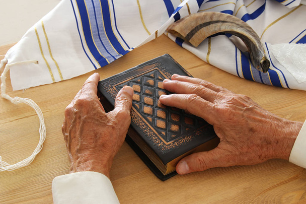 Old Jewish man hands holding a Prayer book, praying, next to tallit and shofar (horn). Jewish traditional symbols. Rosh hashanah (jewish New Year holiday) and Yom kippur concept - Photo, Image