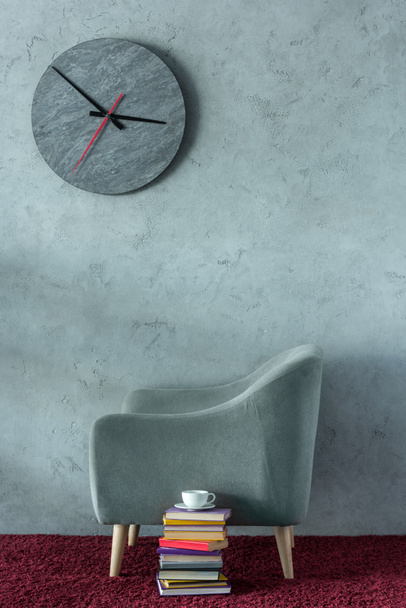 stack of books and cup of coffee near grey armchair in office, clock on wall - Fotografie, Obrázek