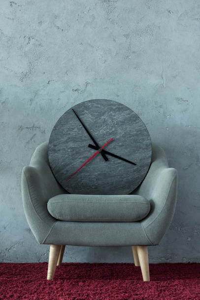 grijze fauteuil met klok op Bourgondische tapijt in de buurt van grijze muur in kantoor - Foto, afbeelding