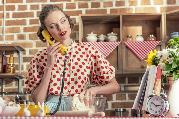 attractive adult housewife talking by phone and kneading dough at kitchen - 写真・画像