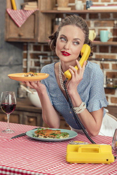 atraente dona de casa adulta com bolo recém-assado falando por telefone rotativo vintage na cozinha
 - Foto, Imagem