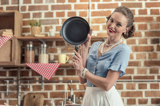 hermosa sonriente ama de casa con sartén mirando a la cámara en la cocina
 - Foto, imagen