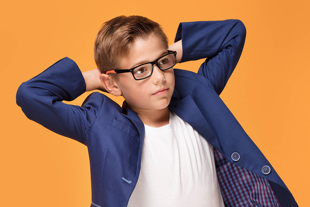 Little elegant boy posing on orange studio backgroud, wearing fashionable eyeglasses and jacket. - Foto, afbeelding
