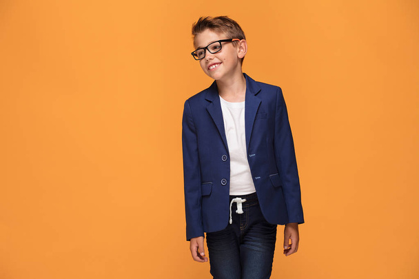 Smiling happy little elegant boy posing on orange studio backgroud, wearing fashionable eyeglasses and jacket. - Φωτογραφία, εικόνα