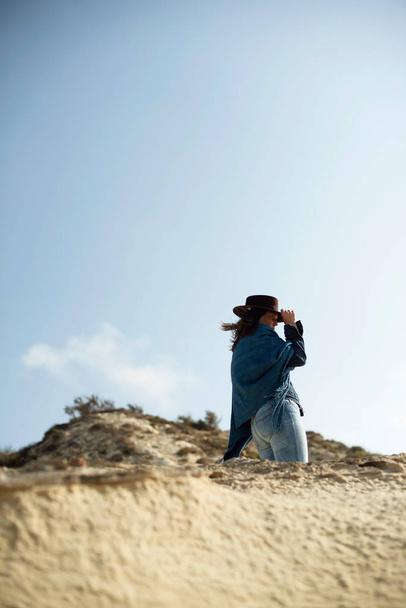 Brunette woman in blue cloth and brown hat standing in desolate windy landscape. - Foto, Imagen