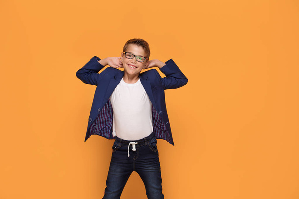 Smiling happy little elegant boy posing on orange studio backgroud, wearing fashionable eyeglasses and jacket. - Photo, image