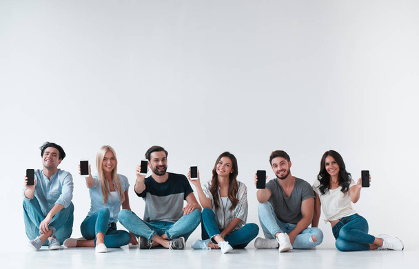 Група молодих людей ізольована на білому тлі. Приваблива молодь сидить разом зі смартфонами в руках. Технологія концепції
. - Фото, зображення
