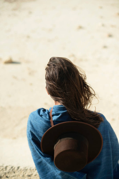 Brunette woman with brown hat in blue cloth standing in windy desert. Rear view. - Foto, afbeelding