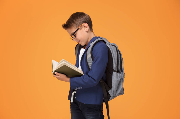 本を保持している小さな学校男の子バックパック オレンジ色背景の上に立って眼鏡で笑顔. - 写真・画像