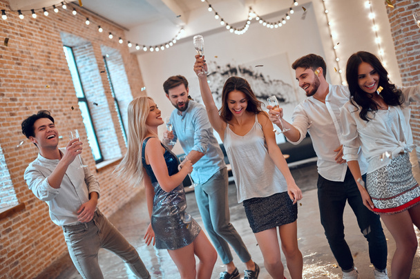 パーティーを始めましょう!一緒に楽しんで若い人たちのグループです。シャンパンと紙吹雪が落ちて大きな明るい部屋で踊る。親しい友人の大きな会社の祝日を祝う. - 写真・画像