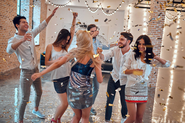 Let the party begin! Group of young people having fun together. Dancing in big light room with champagne and confetti falling. Celebrating holiday in big company of close friends. - 写真・画像