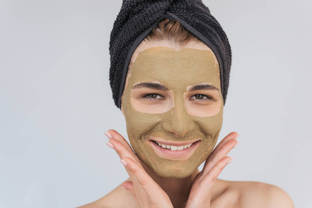 Closeup shot of beautiful young female doing beauty treatment on her face clean skin with clay, looks at the camera with toothy healthy smile, doing wellness. Woman applying organic facial mask. - Фото, изображение