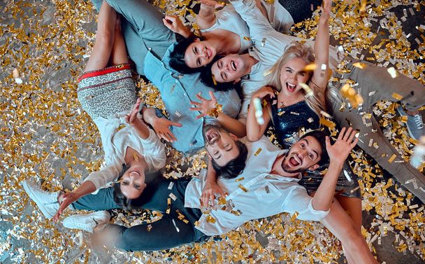 Let the party begin! Group of young people having fun together. Lying on the floor with confetti falling. Celebrating holiday in big company of close friends. - Photo, image