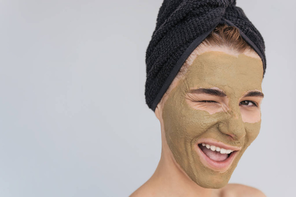 Horizontal studio portrait of happy young woman blink with eye with cosmetic green clay organic mask on her face, wears black towel on hair. Female taking care of face skin, isolated over white wall - Foto, immagini