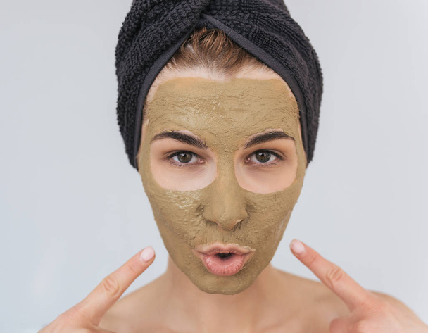 Closeup portrait of happy young woman indicate with fingers on her cosmetic green clay organic mask on her face, wears black towel on hair. Female take care of face skin, isolated on white studio wall - Фото, изображение