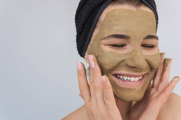 Closeup shot of beautiful young happy woman doing beauty treatment on her face skin. Attractive female smiling with toothy healthy smile and applying facial clay green organic mask, isolated on white - Foto, afbeelding
