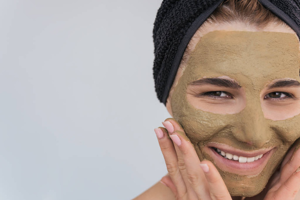 Closeup cropped portrait of beautiful young female doing beauty treatment on her face clean skin with organic clay, looks at the camera with toothy healthy smile. Woman cleansing organic facial mask. - Φωτογραφία, εικόνα