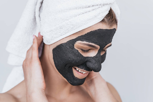 Happy beautiful young woman with black clay face mask doing wellness therapy, posing against white studio wall. Horizontal shot of Caucasian female applying organic facial mask. Beauty, health concept - Фото, изображение