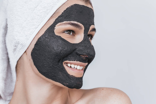 Side closeup view of beautiful smiling young female doing beauty treatment on her face, clean skin with black organic clay, looks aside with toothy healthy smile. Woman applying natural facial mask - Фото, изображение